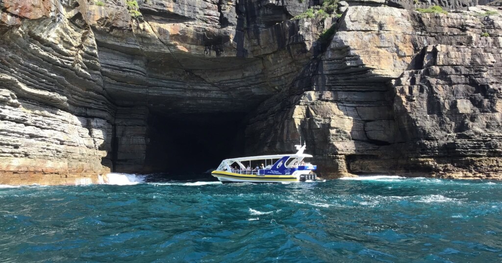 A yellow and white tour boat idling in front of a massive, dark sea cave entrance carved into a towering layered sandstone cliff.