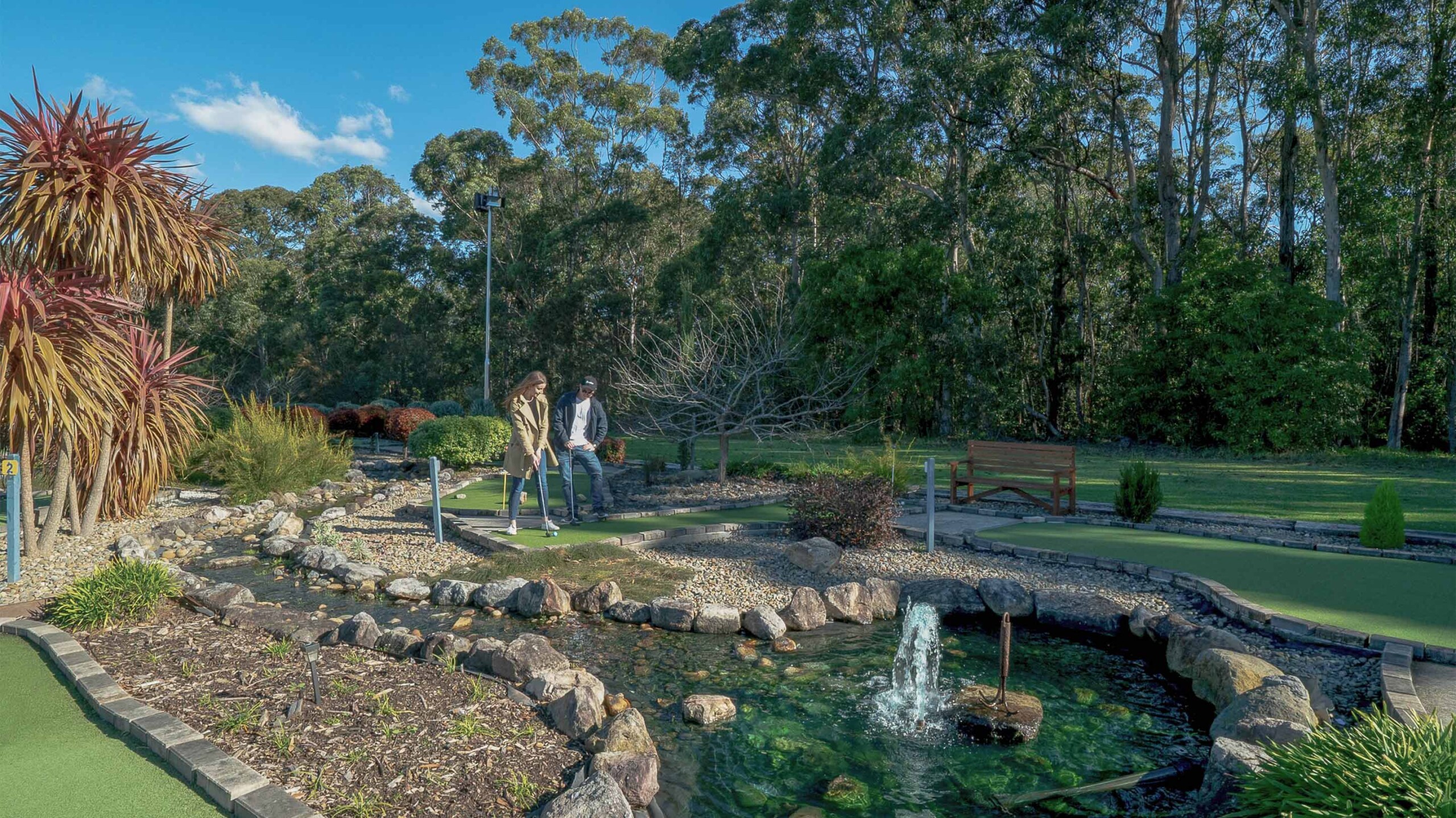 A man and woman playing mini golf on a synthetic green course next to a rock-lined water feature with a small fountain, surrounded by tall eucalyptus trees at Husky Sports.